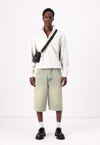 Hanorac gri deschis cu fermoar, pantaloni scurți din denim verde decolorat, geantă crossbody neagră, șosete albe și pantofi masivi negri. Modelul stă cu fața la cameră.