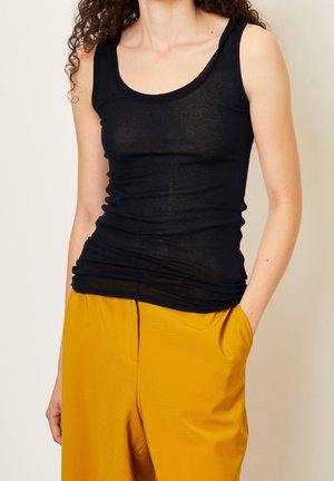 Zwart, nauwsluitend tanktopje van doorschijnende stof met een ronde halslijn en zijplooien, gecombineerd met losse gele broek met een textuurpatroon.