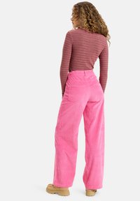 Pantaloni di velluto a coste rosa con vita alta e gambe larghe. Il tessuto presenta costine ben visibili e ci sono tasche a toppa sul retro. Scarpe spesse color beige.
