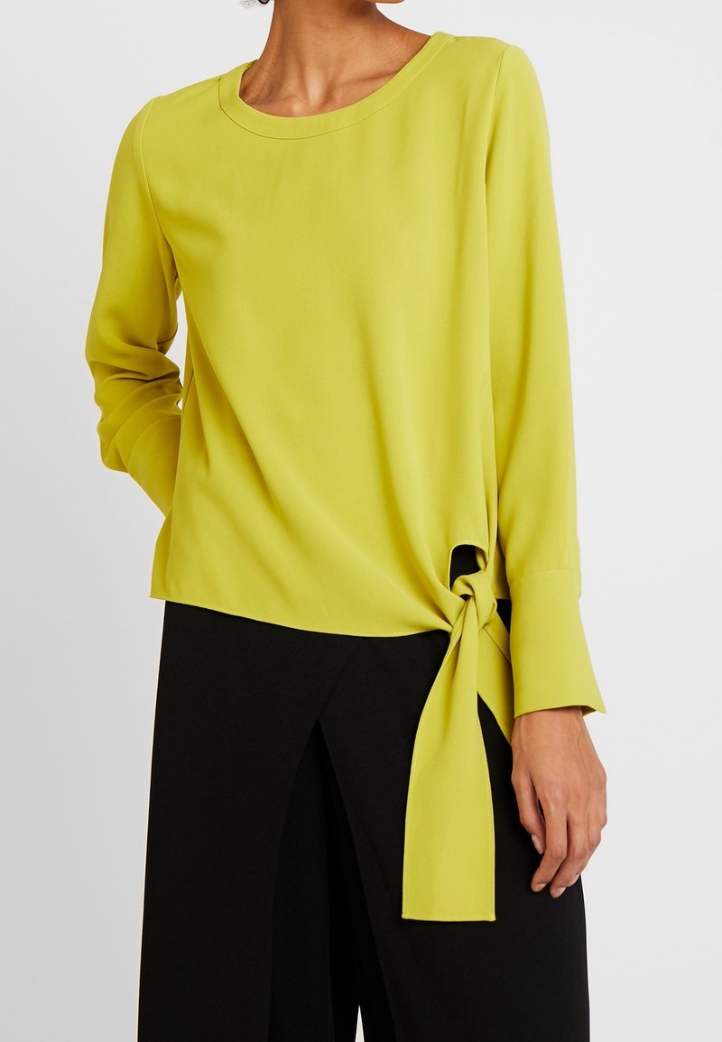 Vihreä pitkähihainen paita, jossa on solmittu yksityiskohta sivussa, pyöreä pääntie ja sileä kangas. Yhdistetty mustiin leveälahkeisiin housuihin.