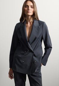 Blazer grigio scuro doppiopetto in tessuto lavorato, con grandi bottoni, rever a punta e una vestibilità ampia. Sono presenti tasche.