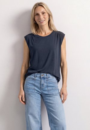 Blonde vrouw staat tegen een witte muur, draagt een mouwloos donkerblauw topje en lichtblauwe high-waisted spijkerbroek met voorkantzakken.