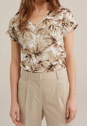 Vrouw draagt beige broek en een top met korte mouwen en V-hals met een bruin-wit abstract bladmotief.
