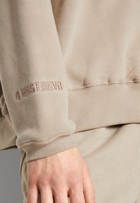 Felpa beige con polsini a costine, caratterizzata da un logo impresso "WRST BHVR" sulla manica. La texture del tessuto è morbida e leggermente strutturata.