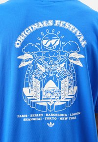 Mėlynas medvilninis marškinėlis su baltu grafiniu dizainu, vaizduojančiu festivalio sceną su palmėmis, garsiakalbiais ir žemiau išvardytomis miestų pavadinimais.