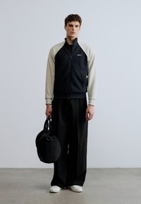 Musta vetoketjullinen takki, jossa on kermaan vivahtavat hihat, yhdistettynä väljiin mustiin housuihin. Malli pitelee pyöreää mustaa laukkua ja käyttää valkoisia kenkiä. Yksinkertainen tausta.