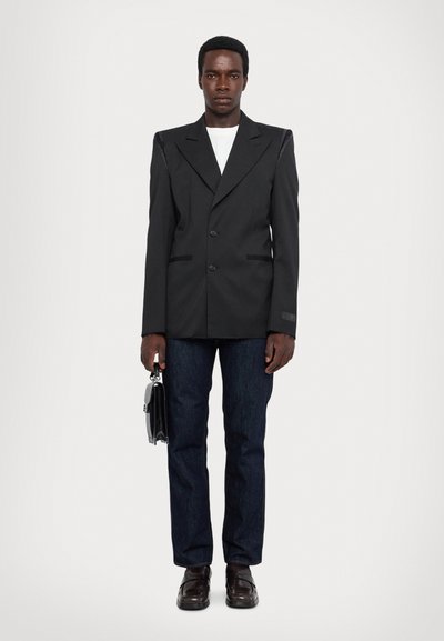 Blazer noir ajusté avec revers en pointe, boutonnières doubles et accents texturés aux épaules, associé à un jean foncé et des chaussures marron. Tenant une mallette noire.