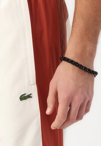 Baldie un sarkanie krāsoti šorti ar zaļu izšūtu krokodila logo. Melna �ķēdes aproce rotā roku.