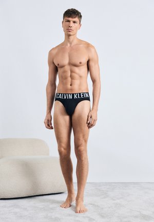 Crne Calvin Klein gaće s širokim sivo-čim logotipom, koje nosi muški model mišićave građe, na neutralnoj pozadini.