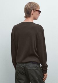 Person mit kurzen blonden Haaren, die Schildkrötenbrillen, einen dunkelbraunen gerippten Pullover und dunkle Jeans trägt, steht nach links vor einem einfarbigen Hintergrund.