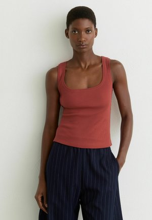 Žena s krátkymi vlasmi, oblečená v bezrukávovom tehlovo sfarbenom topán a námorníckych nohaviciach s jemnými pruhmi, stojaca pred bielou textúrovanou stenou.