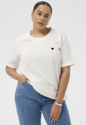 Femeie cu păr închis la culoare, purtând un tricou alb cu o inimă mică neagră pe piept, blugi albaștri și brățară argintie, pozând pe un fundal simplu.