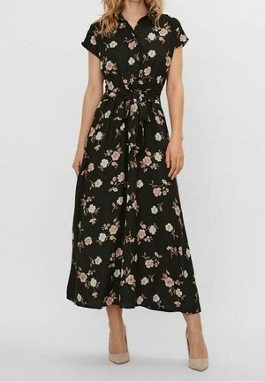 Femme portant une robe midi noire à fleurs avec des manches courtes, un devant boutonné, une taille nouée et des chaussures à talons beiges.