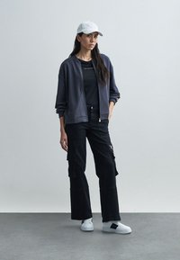 Mørkegrå bomberjakke, svart t-skjorte, svarte cargobukser, hvite joggesko og en lysfarget caps. Avslappet, lagdelt antrekk med funksjonelle lommer.