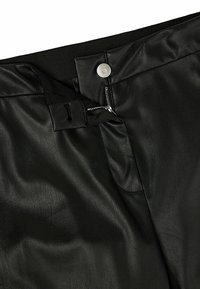Gros plan sur un pantalon en similicuir noir montrant une fermeture éclair partiellement ouverte avec un bouton-pression argenté et une ceinture élastique noire.