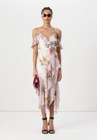 Lichte roze satijnen midi-jurk met ruches over de schouders, een asymmetrische zoom en een getailleerd silhouet. Houdt een rode handtas vast.