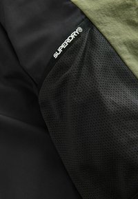 Mustmust ja oliivroheline spordikangas, millel on sile tekstuur ja võrgu sarnane detail. Varrukal on valge "SUPERDRY" logo.