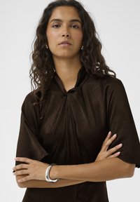 Haut marron texturé avec un col montant et des manches courtes évasées. Détail noué au niveau du col. Bracelet manchette argenté visible.