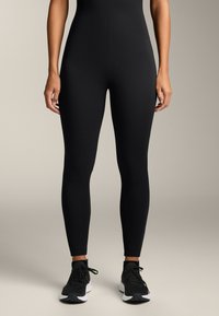 Črno dolgi rokav bodi s prilagojenim krojem, narejen iz gladkega materiala, v kombinaciji s črnimi športnimi čevlji z belimi podplati in vezalkami.