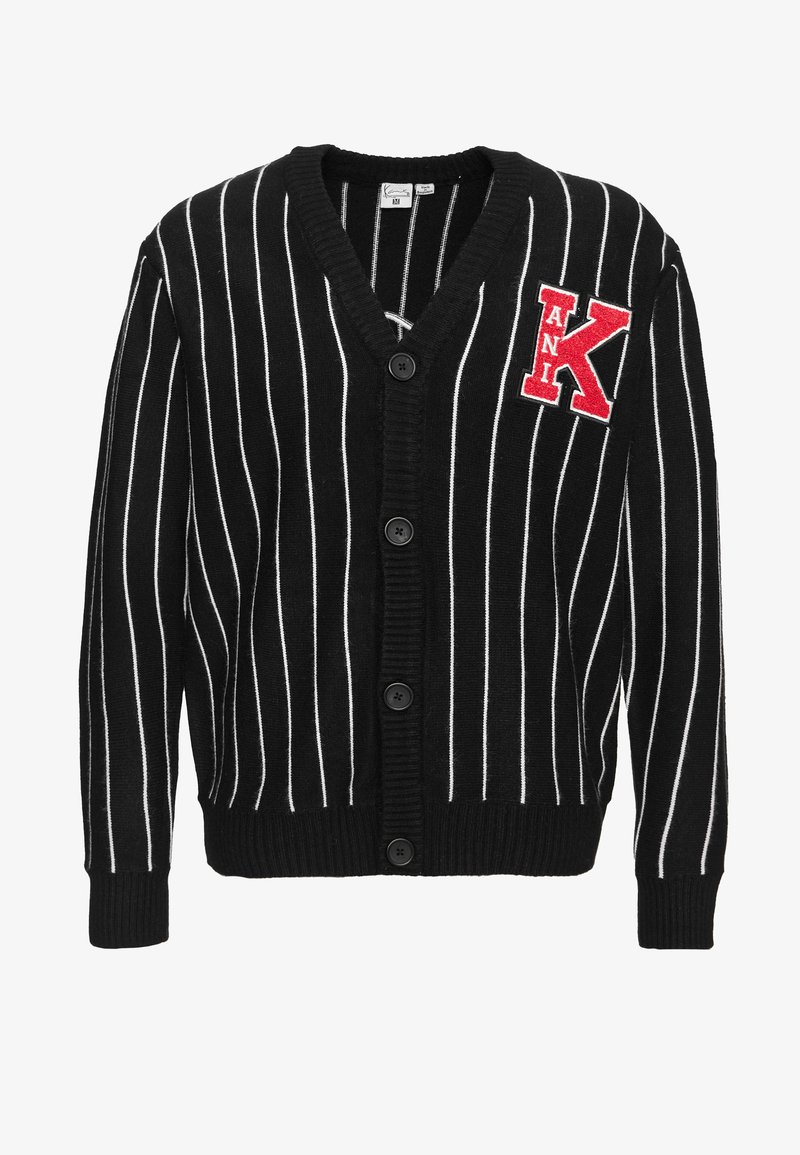 Musta-valkoraidallinen neuletakki V-kaula-aukkolla, nappikiinnityksellä ja punaisella kirjotulla "K"-merkillä. Pehmeä kangas, jossa resorireunaiset hihansuut ja helma.