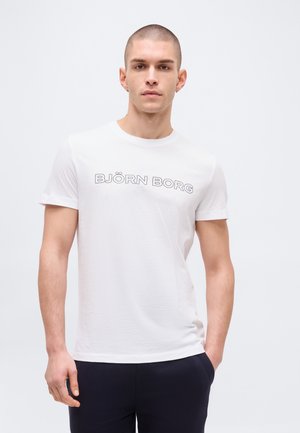 Νεαρός άνδρας με κοντά μαλλιά, φοράει λευκό μπλουζάκι Björn Borg και σκούρο παντελόνι, στέκεται μπροστά σε απλό φωτεινό φόντο.