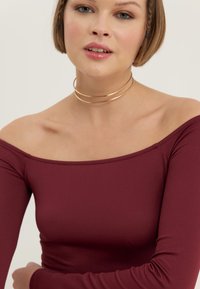 Bordo bluzon bez naramenica s dugim rukavima, napravljen od glatkog materijala. Ima široki izrez i uski siluetu. Zlati choker kao naglasak.