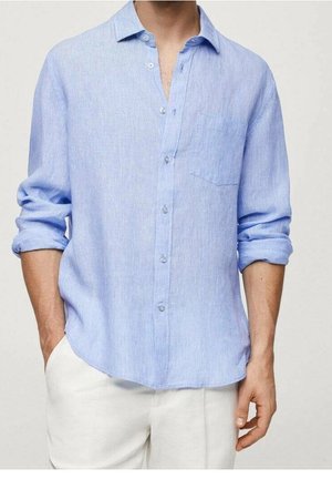 Chemise bleu clair à manches longues avec poche poitrine, portée avec les manches retroussées et assortie à un pantalon blanc. Mains partiellement dans les poches.
