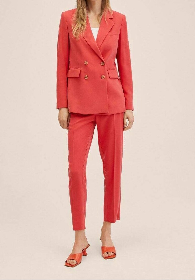 Korallenfarbener, doppelreihiger Blazer mit goldenen Knöpfen, kombiniert mit passenden, schmal geschnittenen Hosen und orangefarbenen Absatzsandalen. Glattes Material, maßgeschneiderter Schnitt.