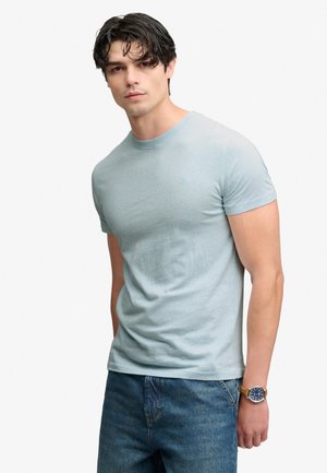 Ung mand med mørkt hår iført en lyseblå t-shirt med korte ærmer, blå jeans og et armbåndsur med brun rem.