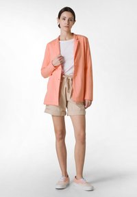 Korallfärgad kavaj, vit t-shirt, beiga shorts och ljusrosa sneakers. Kavajen har knappar och en avslappnad passform; shortsen har uppvikta muddar.
