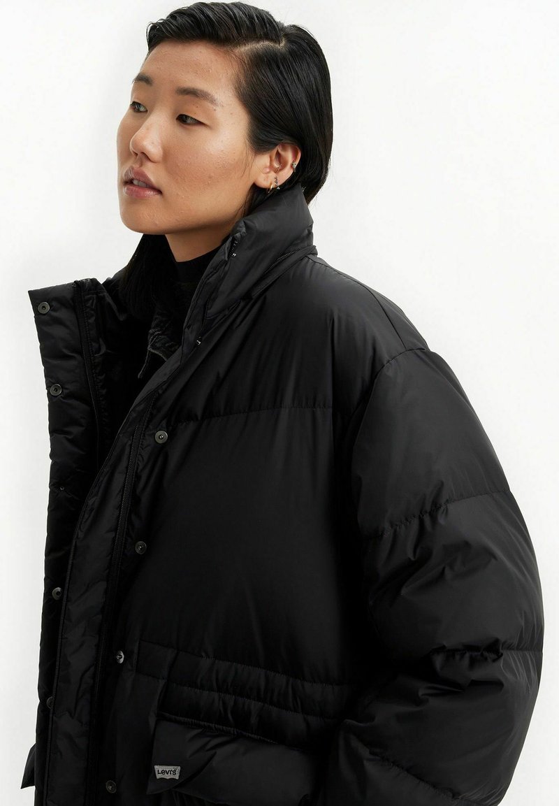 Schwarze Steppjacke mit hohem Kragen, glänzender Oberfläche, Druckknöpfen und einer Fronttasche. Texturierter Stoff mit gepolstertem Design.