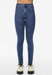 Υψηλής μέσης μπλε τζιν σε ελαστικό υλικό τζιν, με μπροστινές τσέπες και κλασικό σχέδιο πέντε τσεπών, συνδυασμένα με μπεζ παπούτσια.