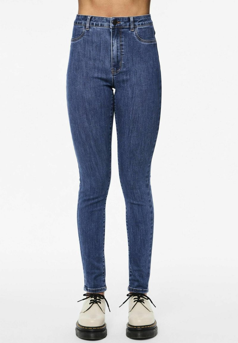 Υψηλής μέσης μπλε τζιν σε ελαστικό υλικό τζιν, με μπροστινές τσέπες και κλασικό σχέδιο πέντε τσεπών, συνδυασμένα με μπεζ παπούτσια.