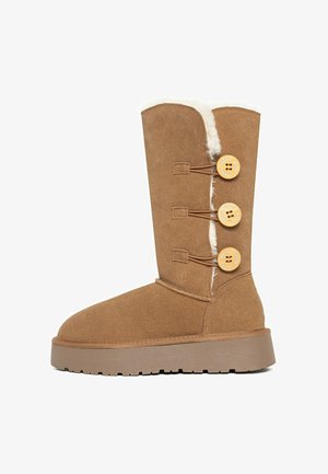 Brauner Wildlederstiefel mit Kunstfell-Futter, drei dekorativen Holzknöpfen und einer klobigen Sohle. Besitzt eine abgerundete Zehenform und eine Höhe bis zur Wade.