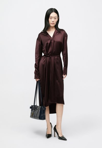 Φόρεμα από βουργουνδί σατέν με V-λαιμόκοψη και ζώνη στη μέση, με μακριά μανίκια και πλευρικό σκίσιμο. Συνδυασμένο με μπλε τσάντα χειρός και μαύρες γόβες.