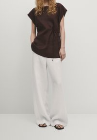 Tunik hitam coklat dengan lengan pendek dan ikatan samping, dipasangkan dengan celana linen abu-abu muda yang lebar dan sandal hitam dengan aksen melingkar.