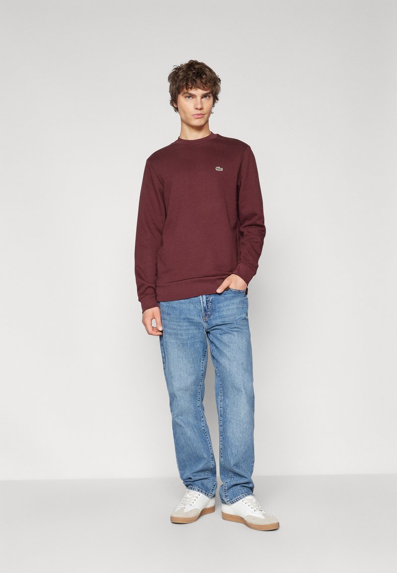 Bordowy sweter z okrągłym dekoltem, ściągaczami przy rękawach i u dołu. W zestawie z jasnoniebieskimi dżinsami i białymi trampkami z beżowymi akcentami.