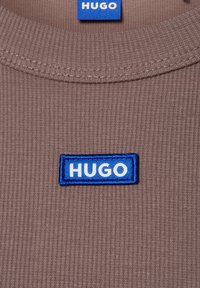 Brązowy t-shirt z żebrowanej tkaniny z niebieską prostokątną haftowaną metką z białym napisem "HUGO" na dekolcie.