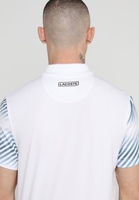 Vit polo-tröja med blå och vita randiga korta ärmar, med krage och Lacoste-logotyp på baksidan av nacken. Slät textur.