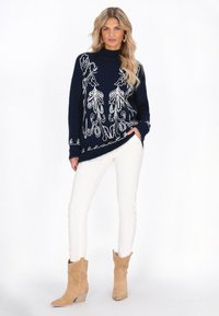 Pulover tricotat bleumarin cu broderie florală albă, guler înalt și fante laterale. Asortat cu pantaloni albi ajustați și cizme scurte bej.