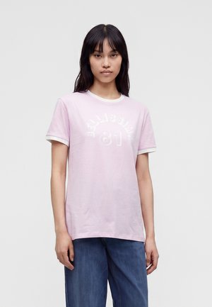 Joven mujer con cabello largo y oscuro lleva una camiseta rosa claro con ribetes blancos y texto "BELLISSIMA 81", combinada con jeans azules.