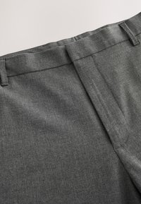 Gros plan d’un pantalon habillé gris foncé montrant la ceinture, les passants de ceinture et les détails de la couture avant sur un fond clair uni.