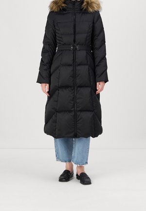 Manteau d'hiver long noir matelassé avec capuche bordée de fourrure, taille ceinturée, porté sur un jean bleu clair raccourci et des mocassins noirs.