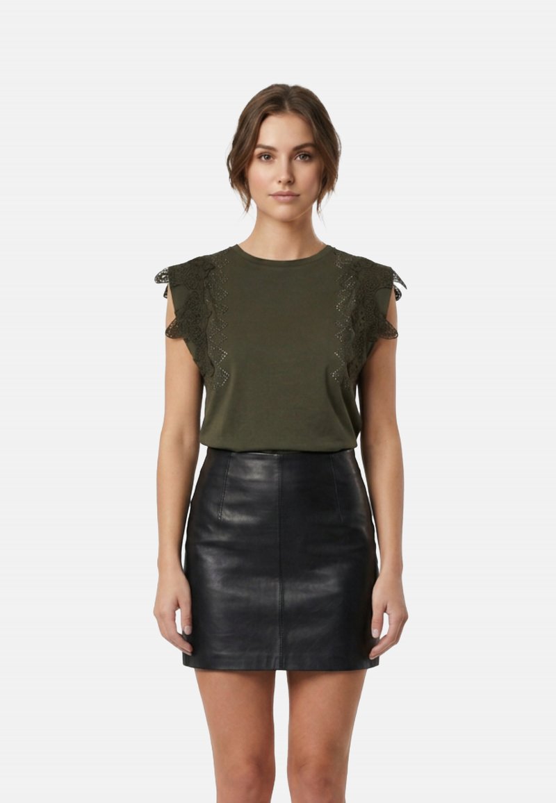 Frau trägt ein olivgrünes, ärmelloses Oberteil mit Spitzenvolants und einen schwarzen Leder-Minirock, steht frontal vor einem schlichten Hintergrund.