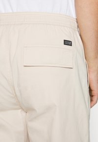 Beige elastische Bundhose mit einer Gesäßtasche und einem schwarzen Stoffetikett. Die Textur wirkt glatt und leicht.