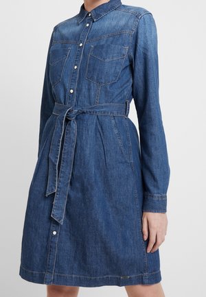 Zbliżenie na osobę ubraną w niebieską sukienkę koszulową z denimu z przednimi kieszeniami, zatrzaskami, kołnierzykiem, długimi rękawami i paskiem wiązanym w talii.