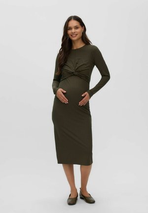 Femme enceinte portant une robe midi à manches longues vert olive avec un détail noué, debout et souriante sur un fond clair uni.