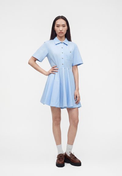 Γυναίκα όρθια με το χέρι στη μέση, φορώντας ανοιχτό μπλε κοντομάνικο φόρεμα με κουμπιά, λευκές κάλτσες και καφέ παπούτσια με πλατφόρμα σε μονόχρωμο φόντο.