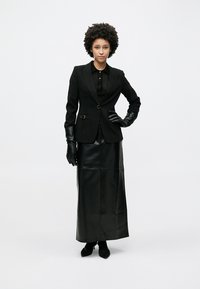 Μαύρο σακάκι ραμμένο στα μέτρα με ένα κουμπί, σε συνδυασμό με μια μακριά μαύρη δερμάτινη φούστα. Το μοντέλο φοράει μαύρα γάντια και μαύρο πουκάμισο με γιακά από κάτω.