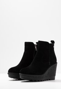 Svarta mockakängor med kilklack och texturerad plattform, sidodragkedja och en dragögla bak. Slät yta med rundad tå.